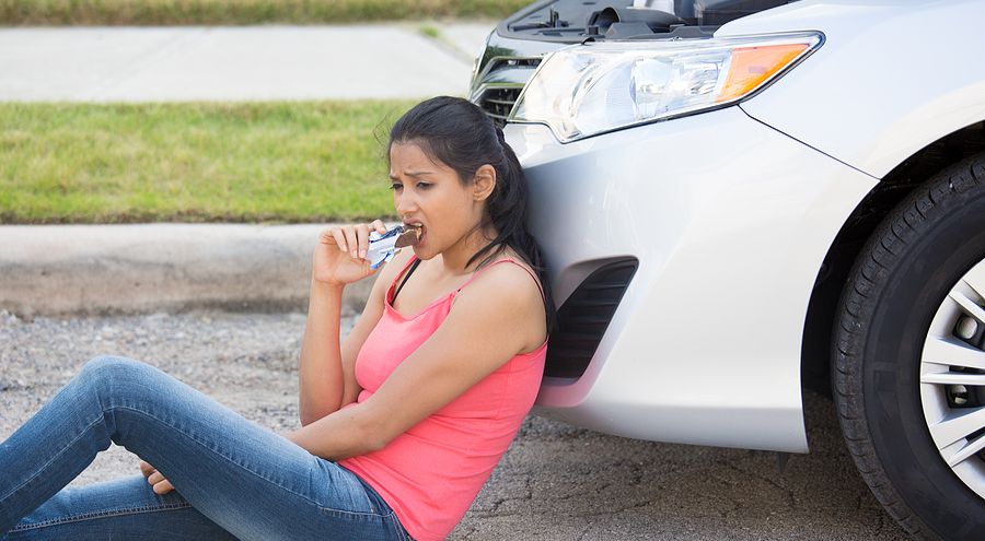 A woman sitting on the ground in front of her car looking depressed and eating a candy bar.