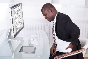 Man sitting at a desk with his hand on his lower back in pain.